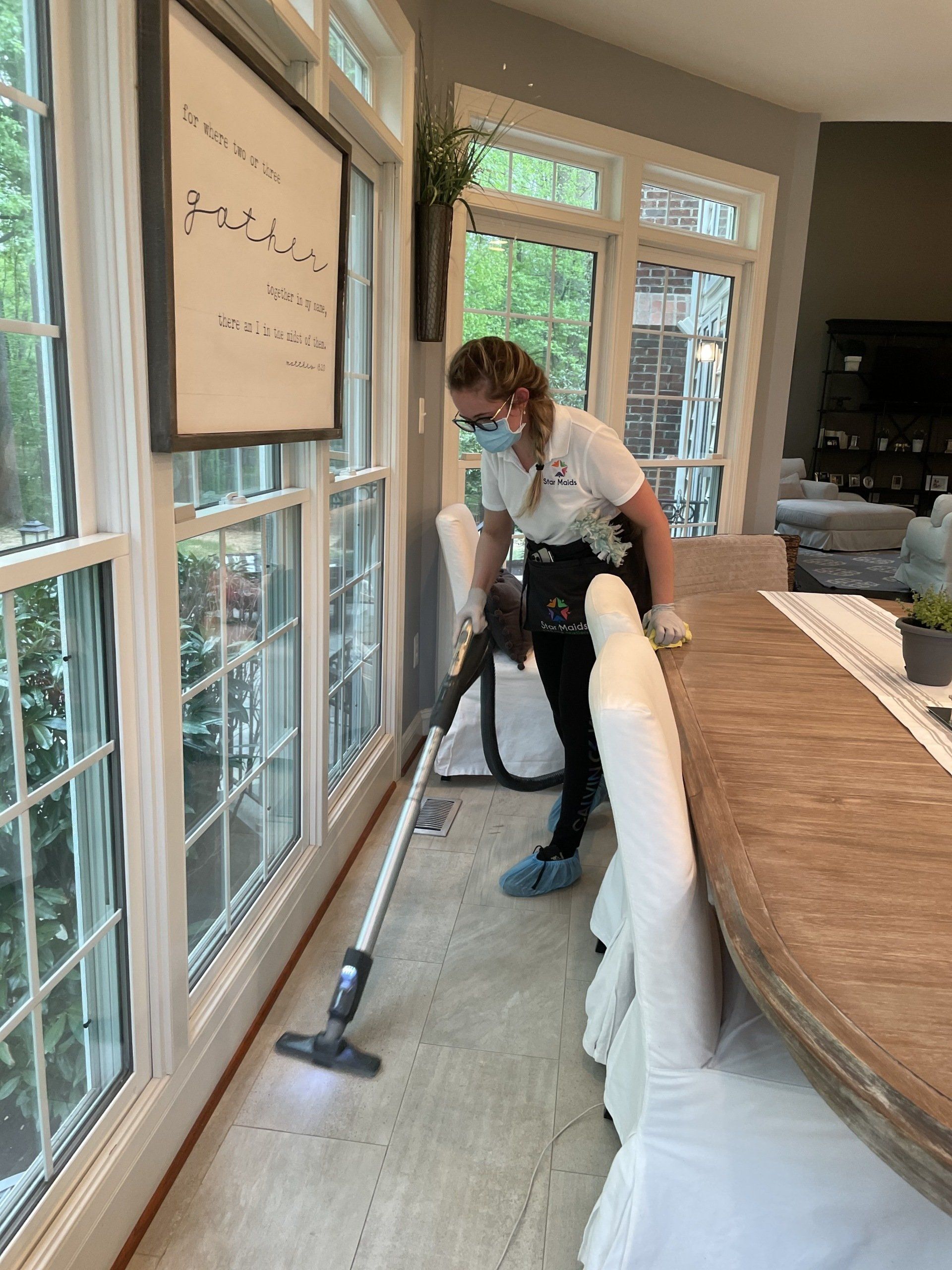 Person vacuuming a bright room beside a long wooden table and large windows