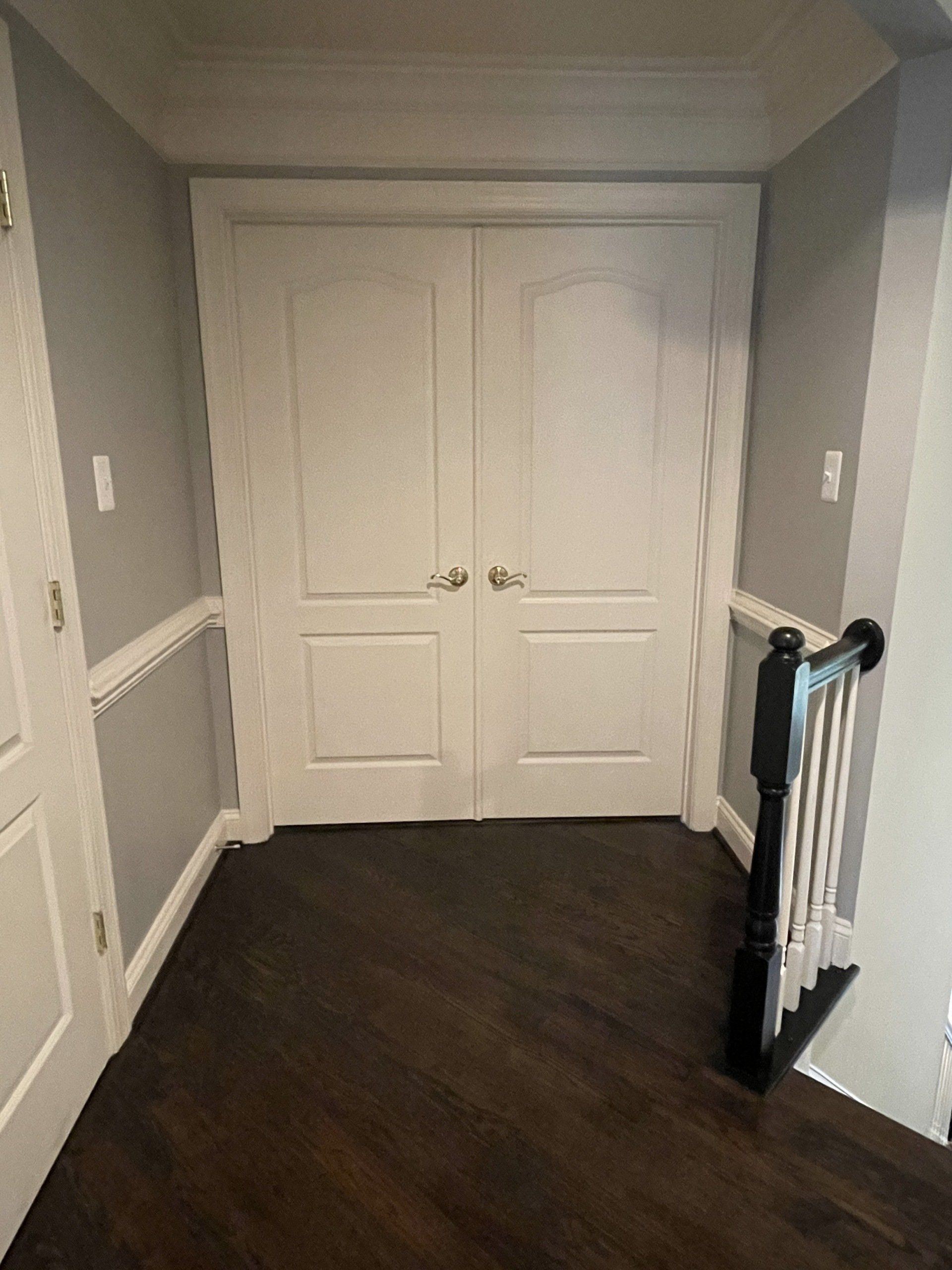 Narrow hallway with white double doors, gray walls, dark wood floor, and stair railing on right.