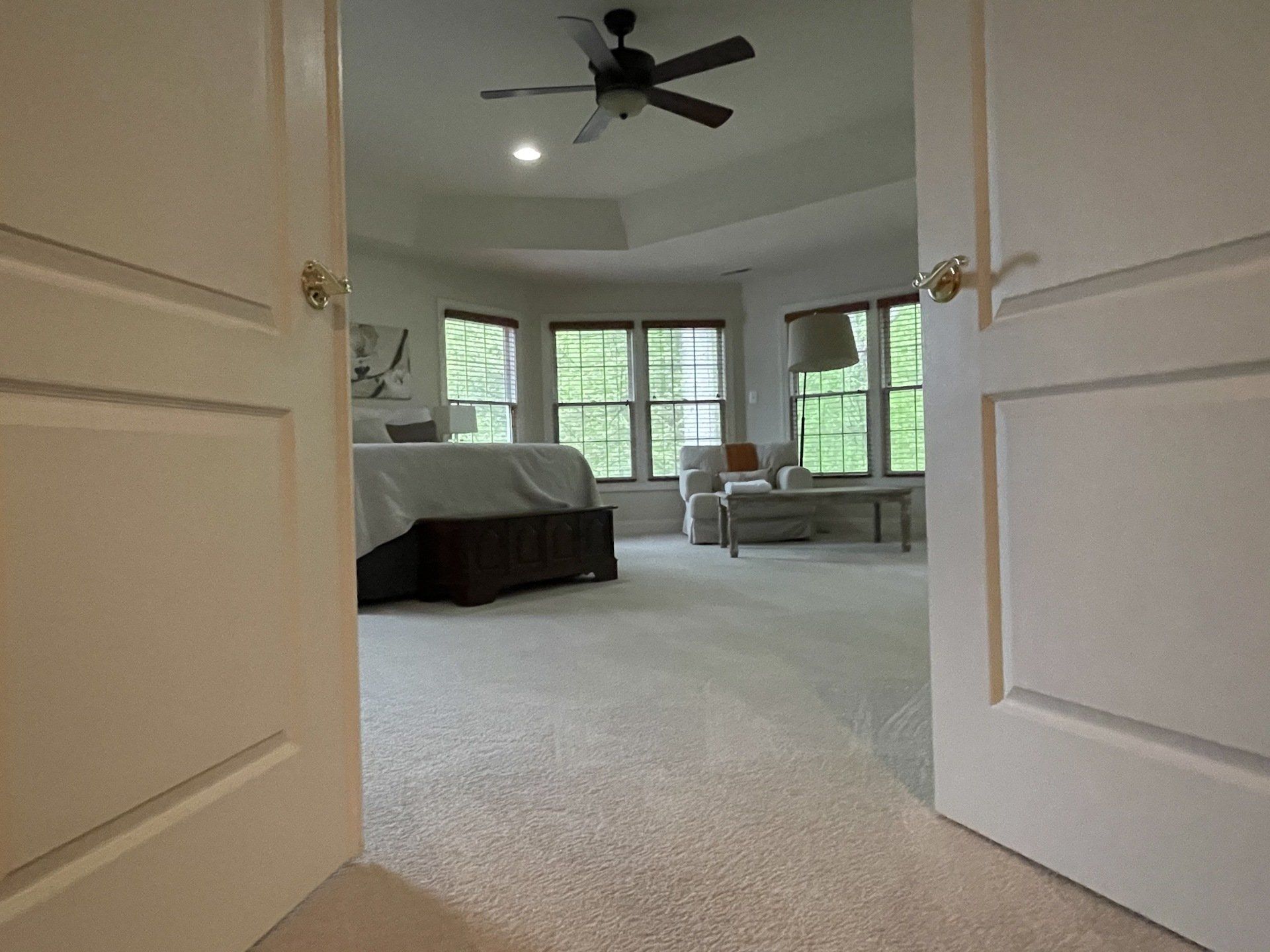 Open double doors to a bright bedroom with a bed, large windows, and a ceiling fan.