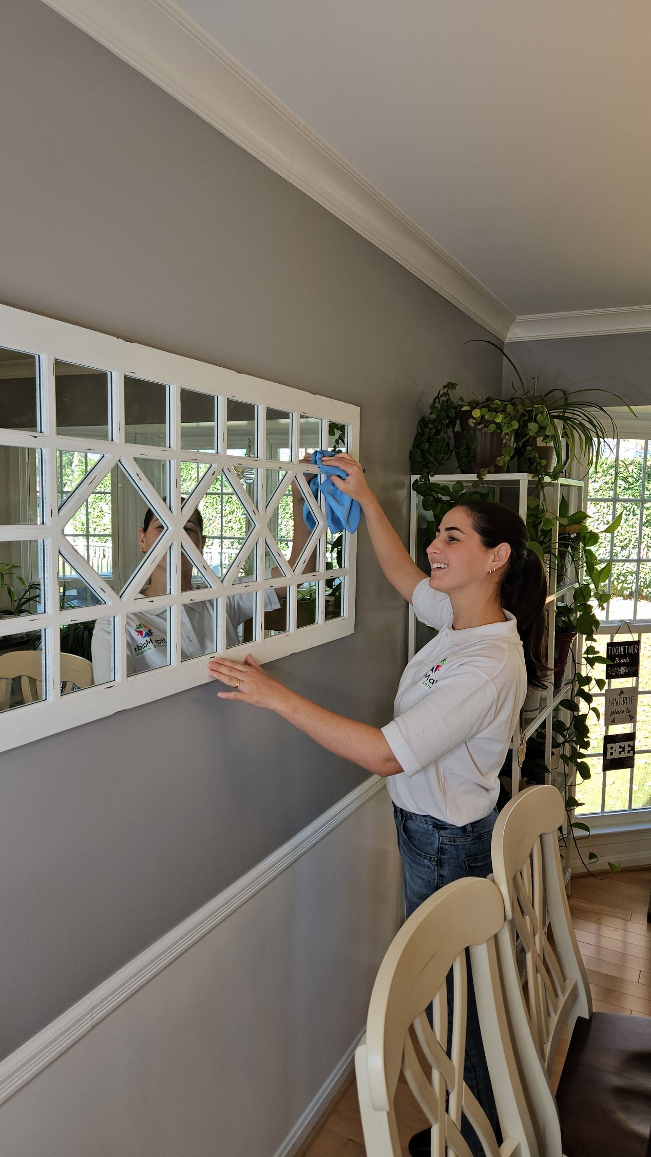 Person dusts a gray wall mirror with a blue cloth in a bright room with plants and chairs