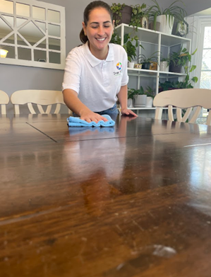 Woman wiping a wooden dining table with a blue cloth in a bright home interior