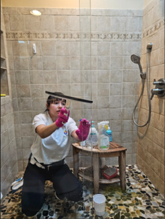 A person wearing pink gloves uses a squeegee to clean a glass shower door inside a tiled shower stall. Person kneeling in a tiled shower, wearing pink gloves and cleaning supplies nearby.