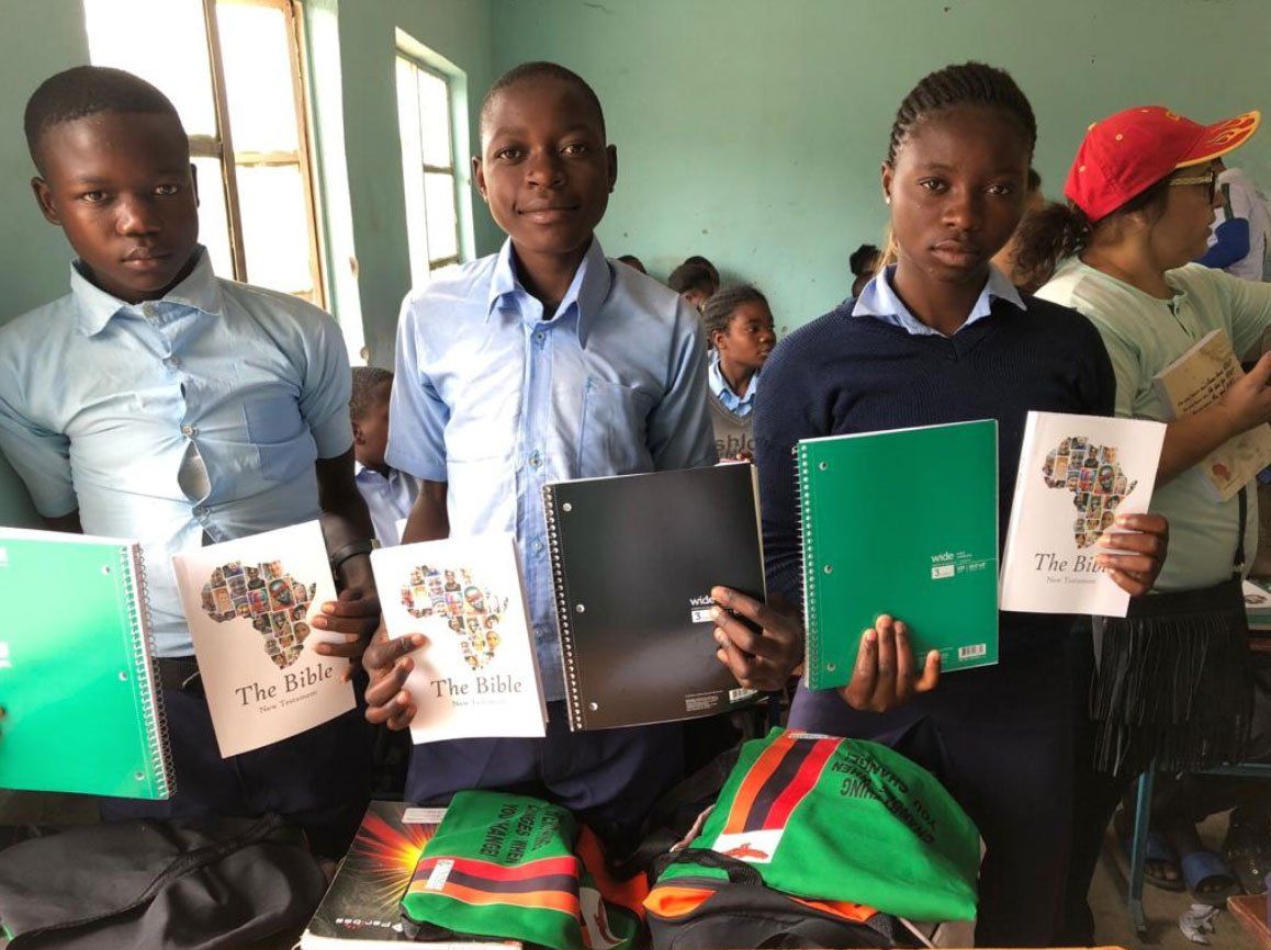Students holding notebooks in a classroom, seated at desks near a window.