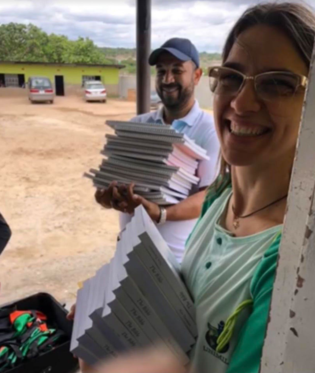 Two smiling people holding stacks of papers on a porch, with parked vehicles and a grassy field behind them.