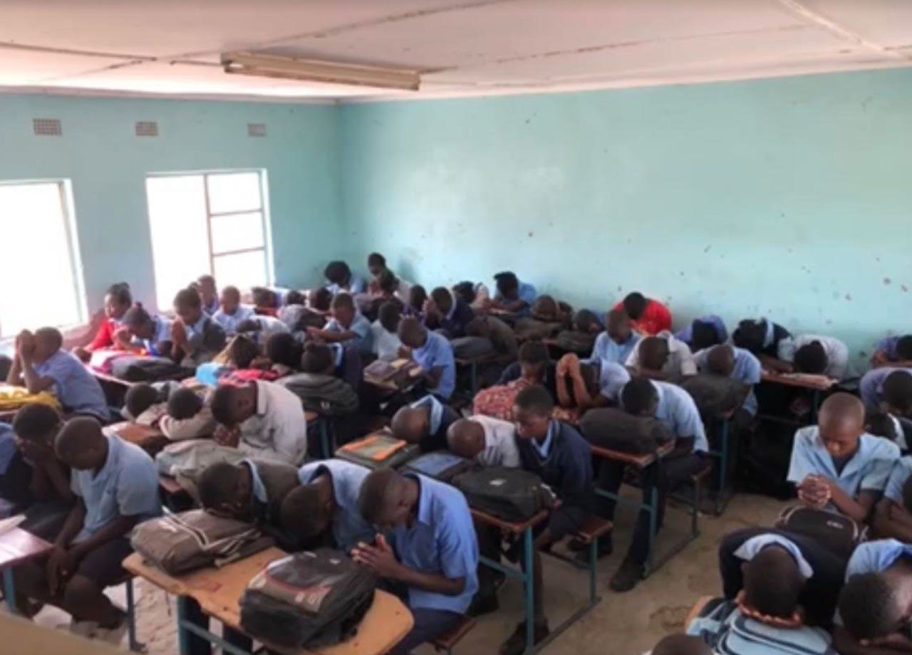 Students taking an exam in a crowded classroom, seated at desks with papers and backpacks.