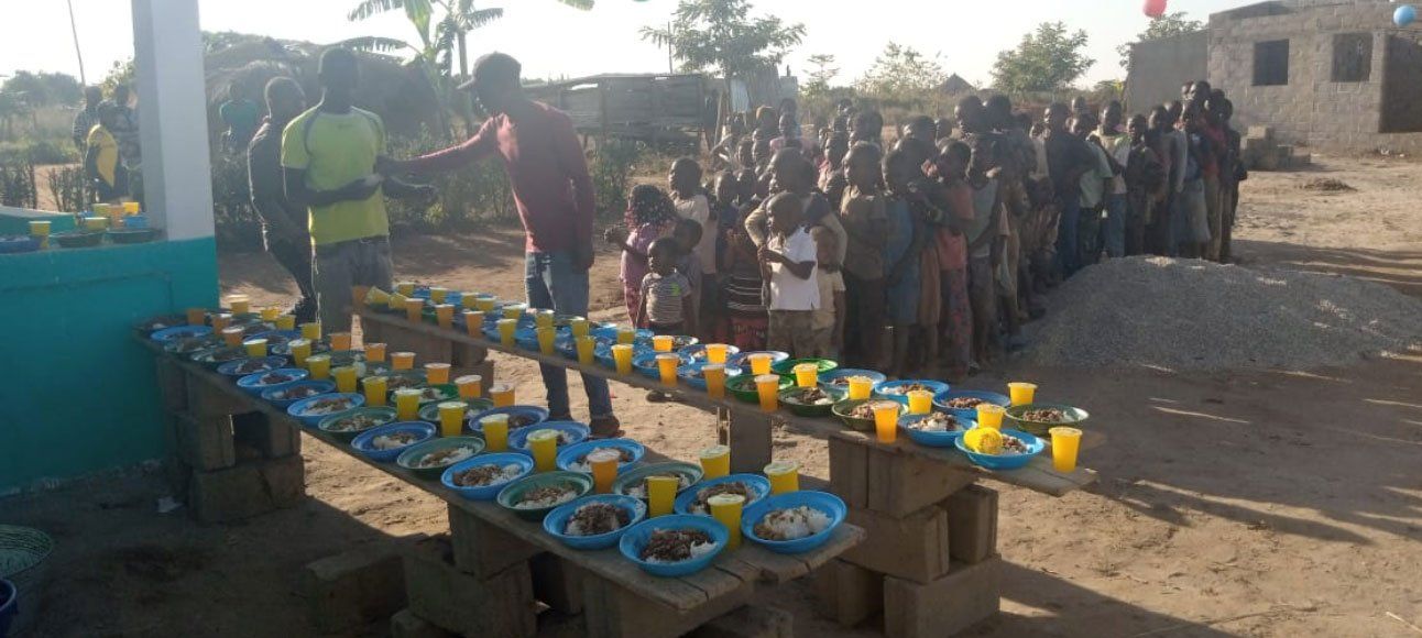 People standing by rows of yellow food containers on tables outdoors at a community gathering