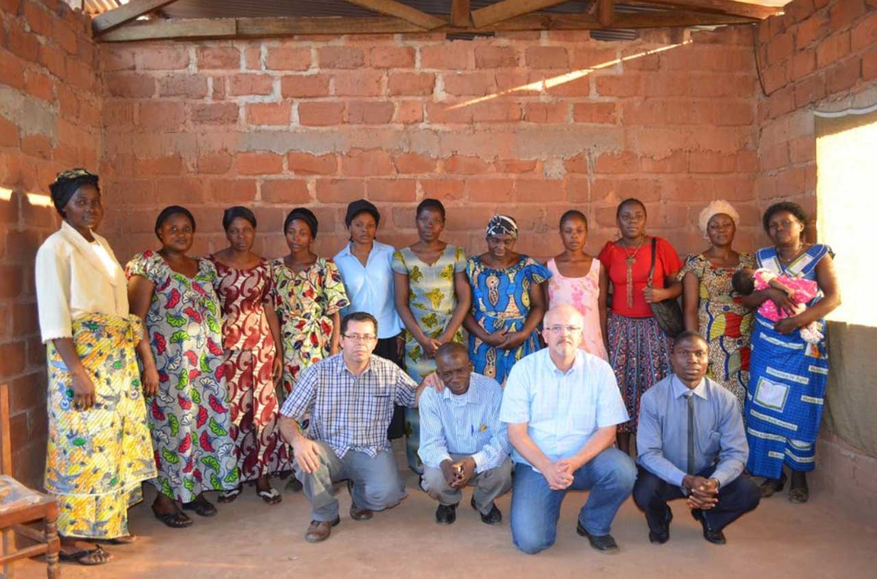 Group of people posing indoors against a brick wall, with several standing and four kneeling in front.
