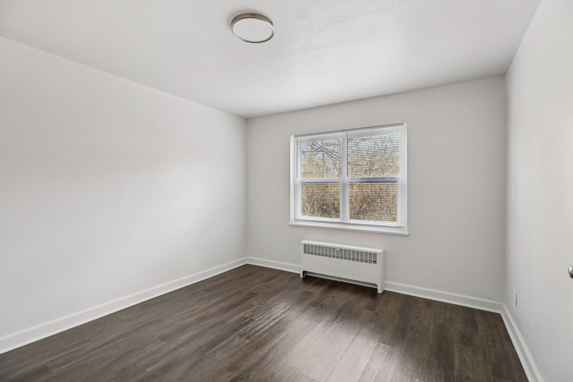 Empty room with hardwood floors, white walls, small window, and radiator.