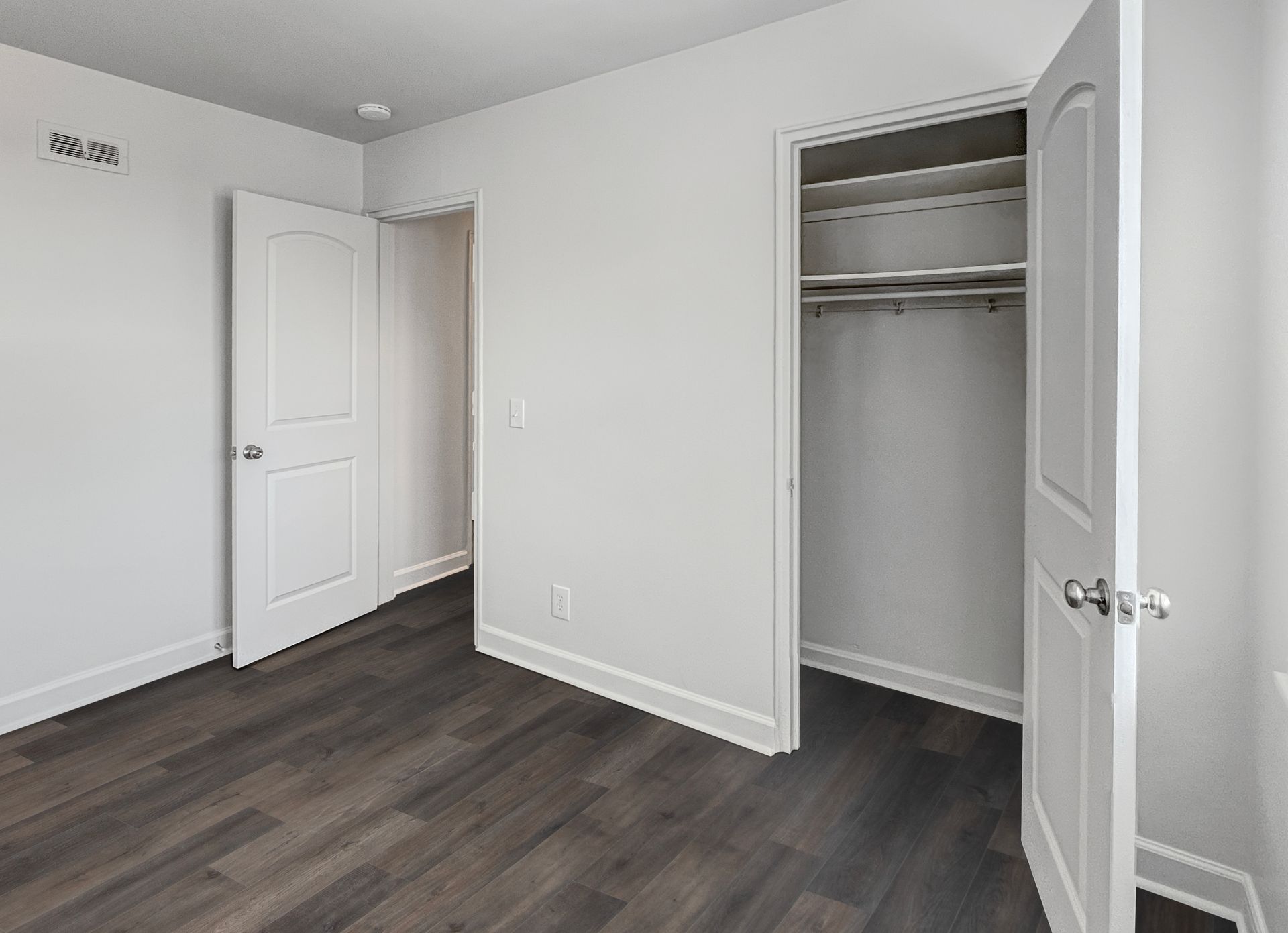 Empty bedroom with dark wood flooring, white walls, and a closet. Two white doors are open.