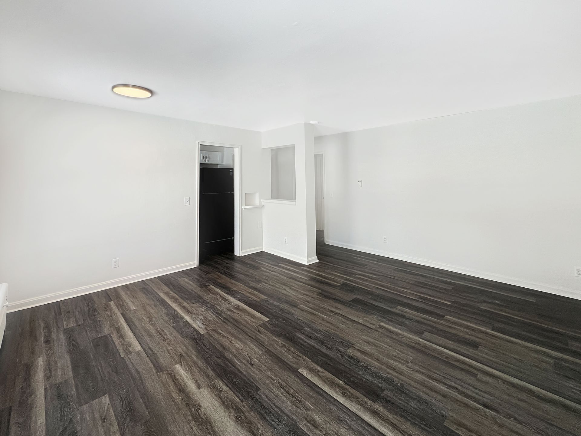 Empty room with dark wood-look flooring, white walls, and a doorway leading to a dark room.
