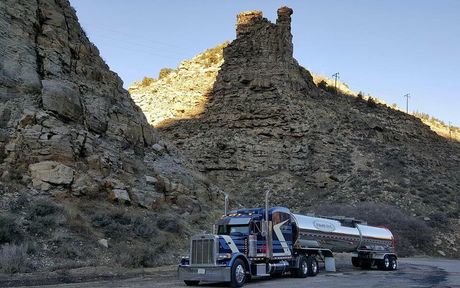 A blue and silver tanker truck parked in a canyon with a large rock formation in the background.