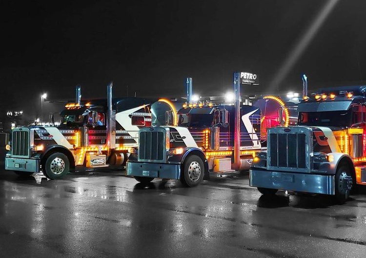 Three illuminated semi-trucks parked at night; colorful lights reflect on wet pavement.