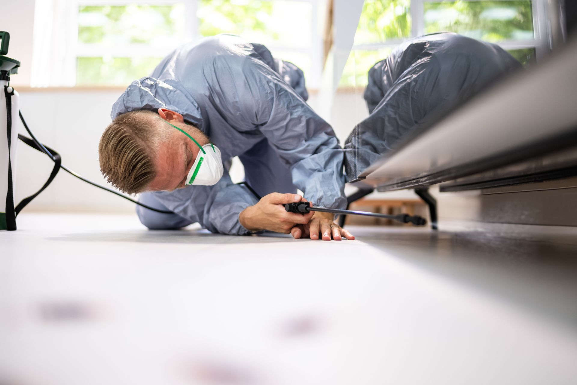 Pest control worker in protective suit spraying under a bed in a room.