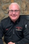 A person with short white hair and glasses, wearing a black shirt with red embroidery. A person with short white hair and glasses, wearing a black shirt with red embroidery.