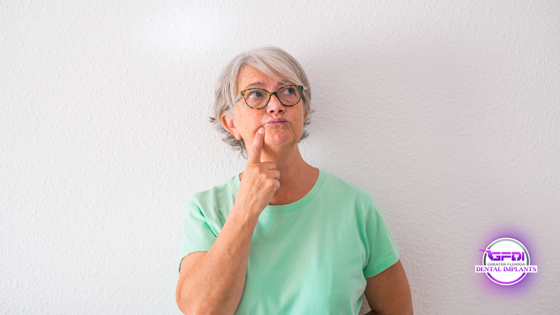 Woman with glasses, gray hair, and a teal shirt, touching chin while looking up thoughtfully against a white wall.