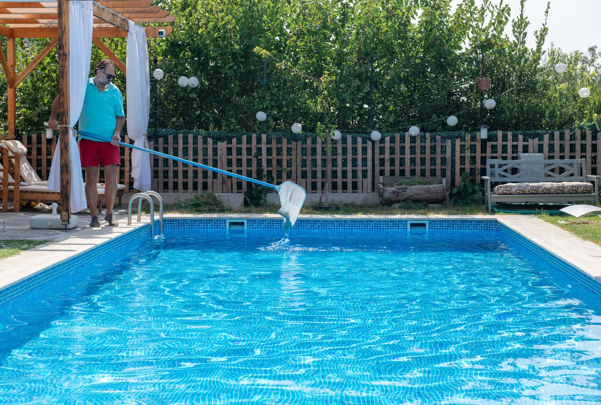 A person in a blue shirt and red shorts cleans a rectangular backyard swimming pool with a long-handled net.