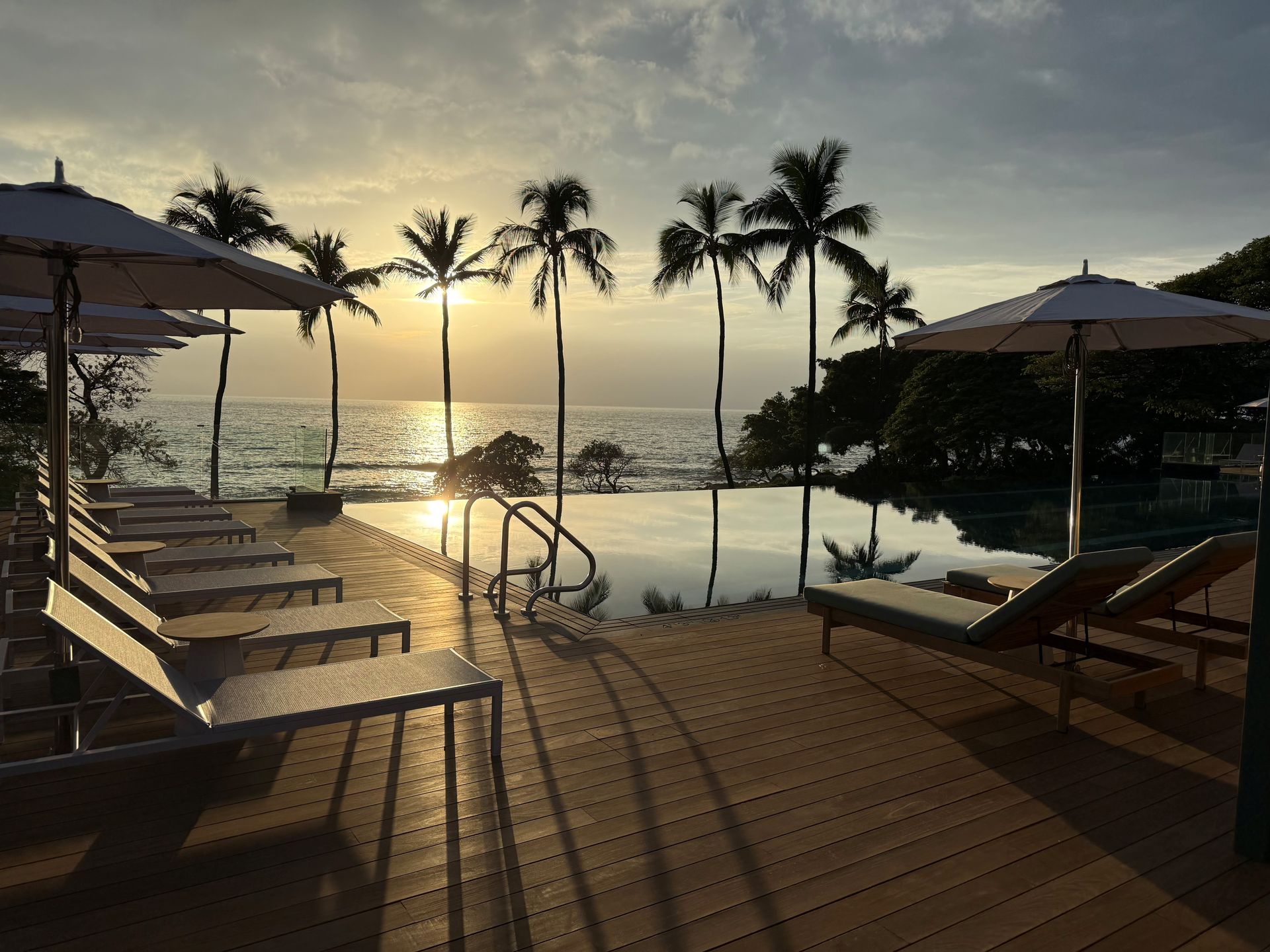 Sunset over a wooden deck with lounge chairs and infinity pool, palm trees, and ocean.