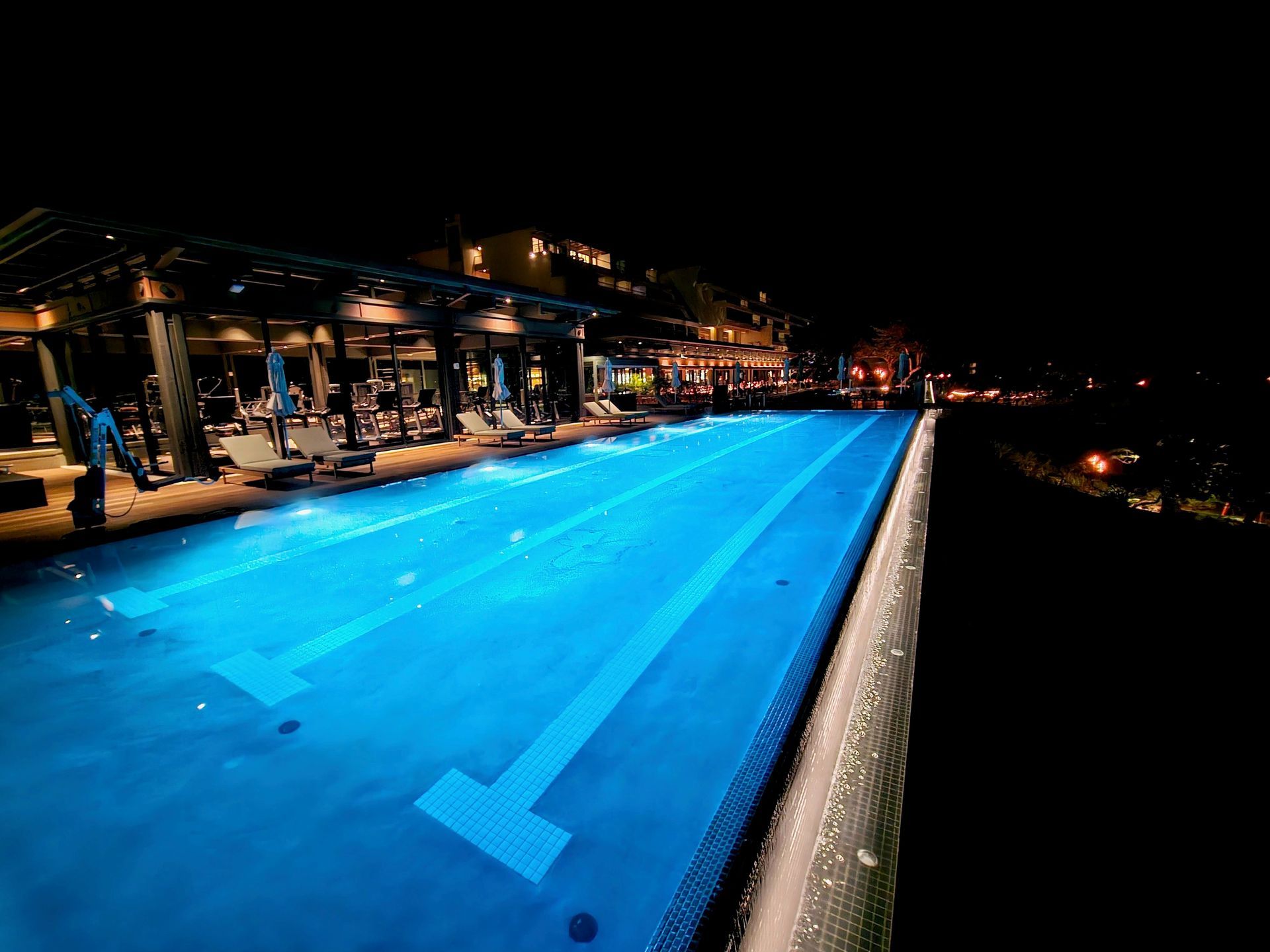 Nighttime view of an illuminated blue swimming pool next to a building with outdoor seating; dark background.