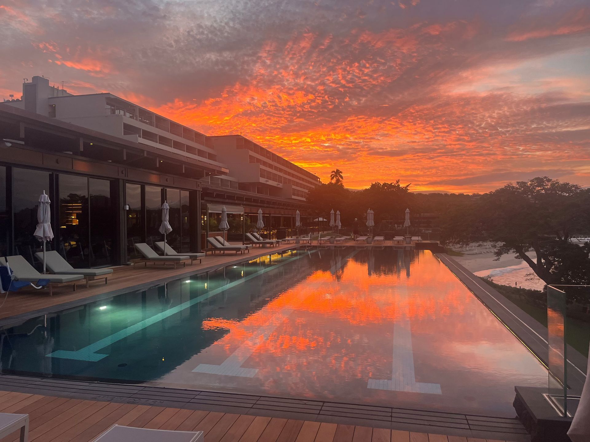 Pool reflecting fiery sunset over a modern building with lounge chairs.