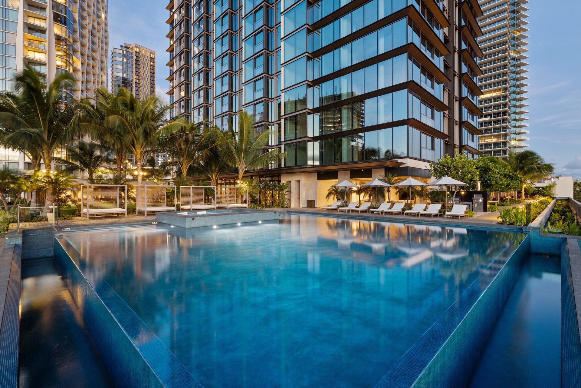 Swimming pool with lounge chairs, palm trees, and modern high-rise buildings.