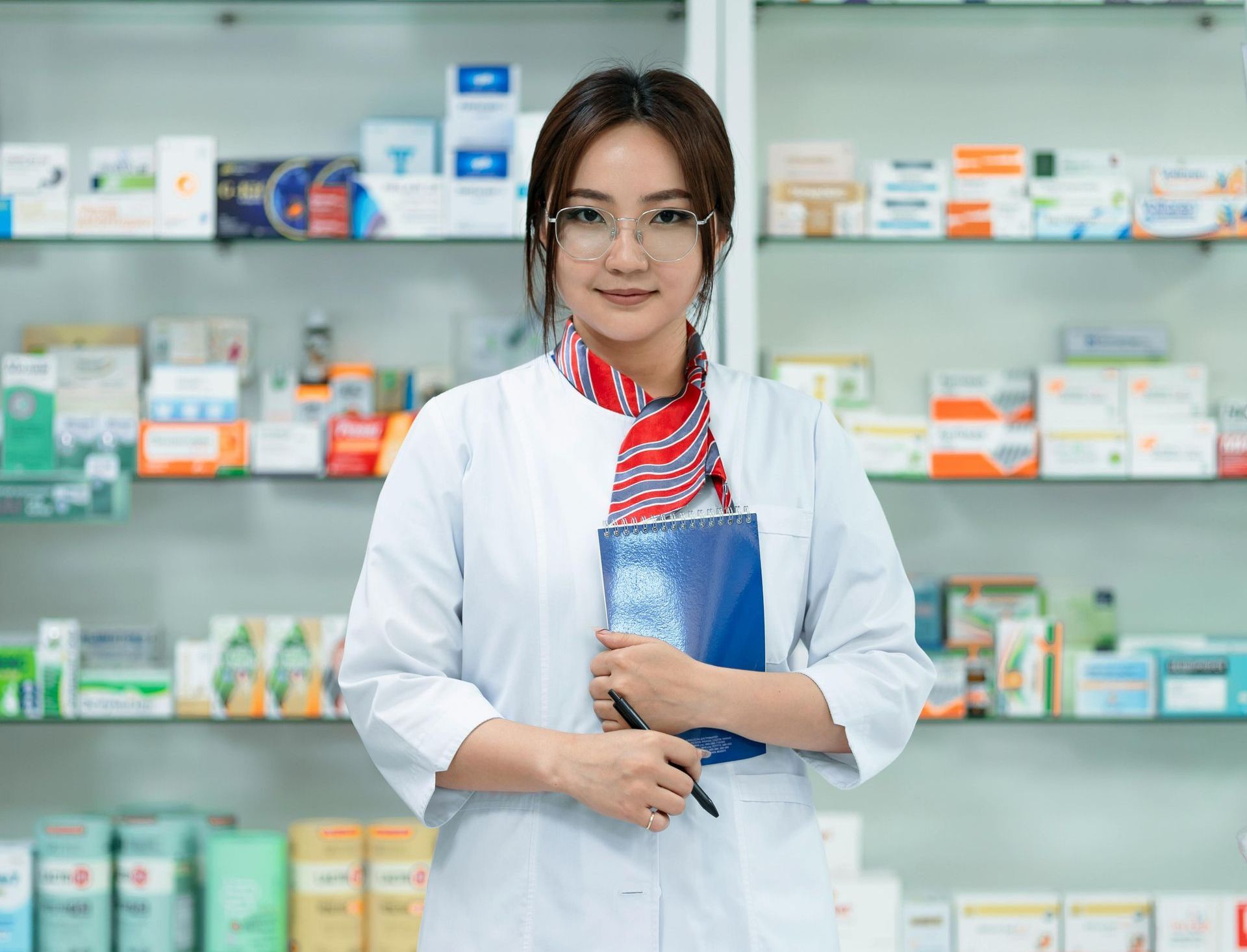 Pharmacist in white coat stands in front of shelves of medications, holding a blue pad, smiling.