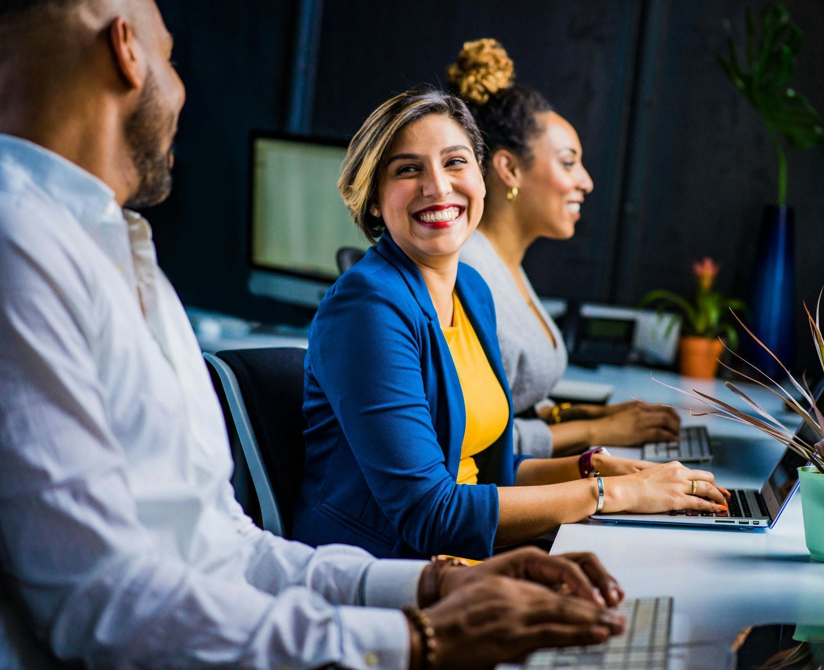 Three diverse coworkers smiling and working at desks in an office.