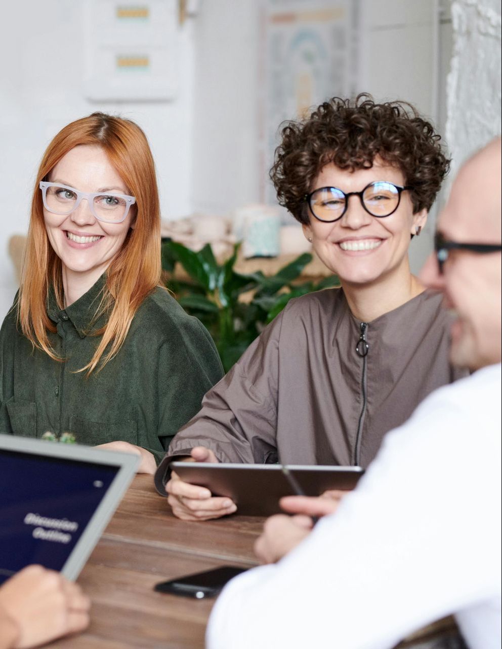 Two smiling women in glasses at a table, one red-haired, one with curly hair. A man is partially visible.