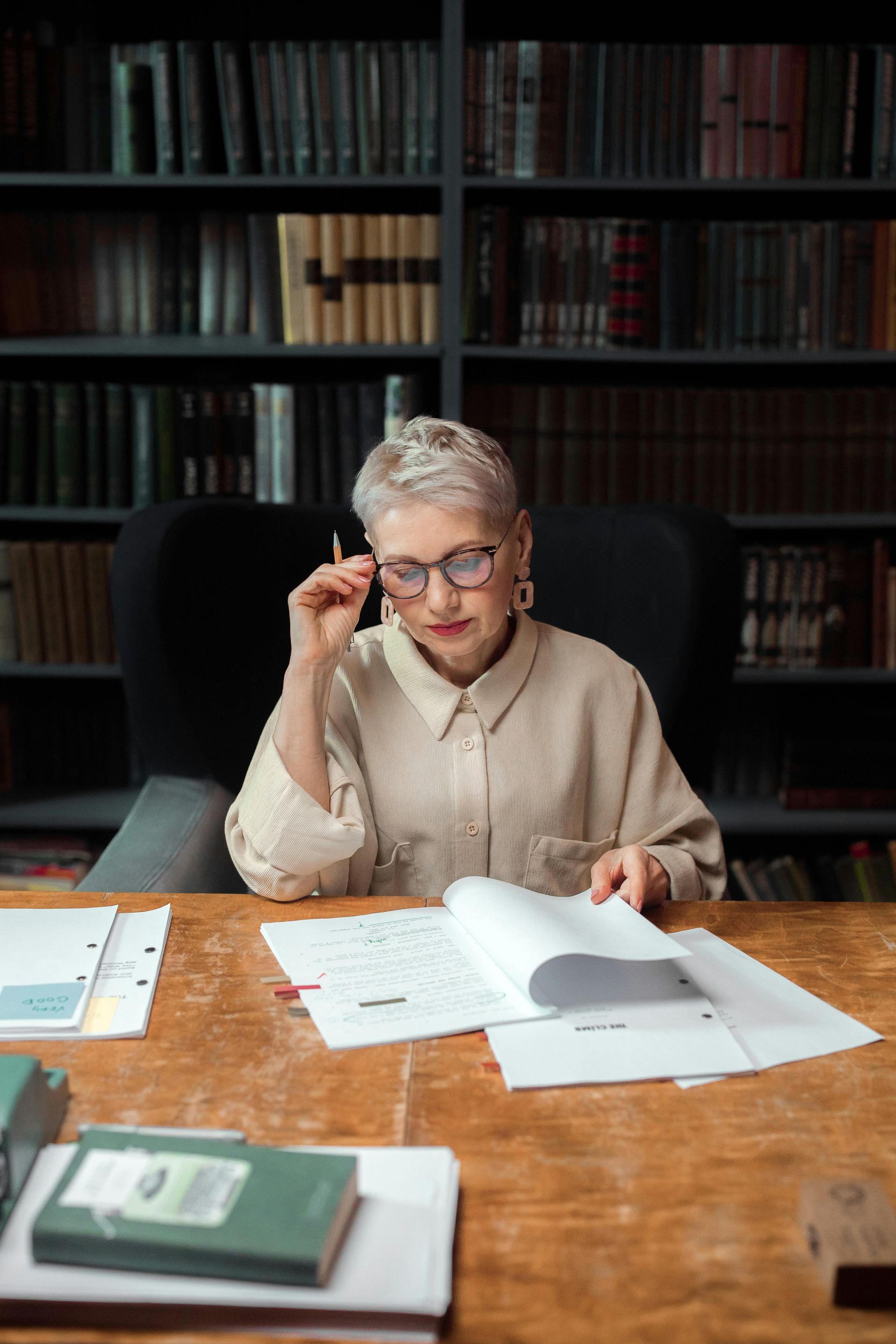 Woman with short gray hair, wearing glasses, reading documents at a desk in a library.