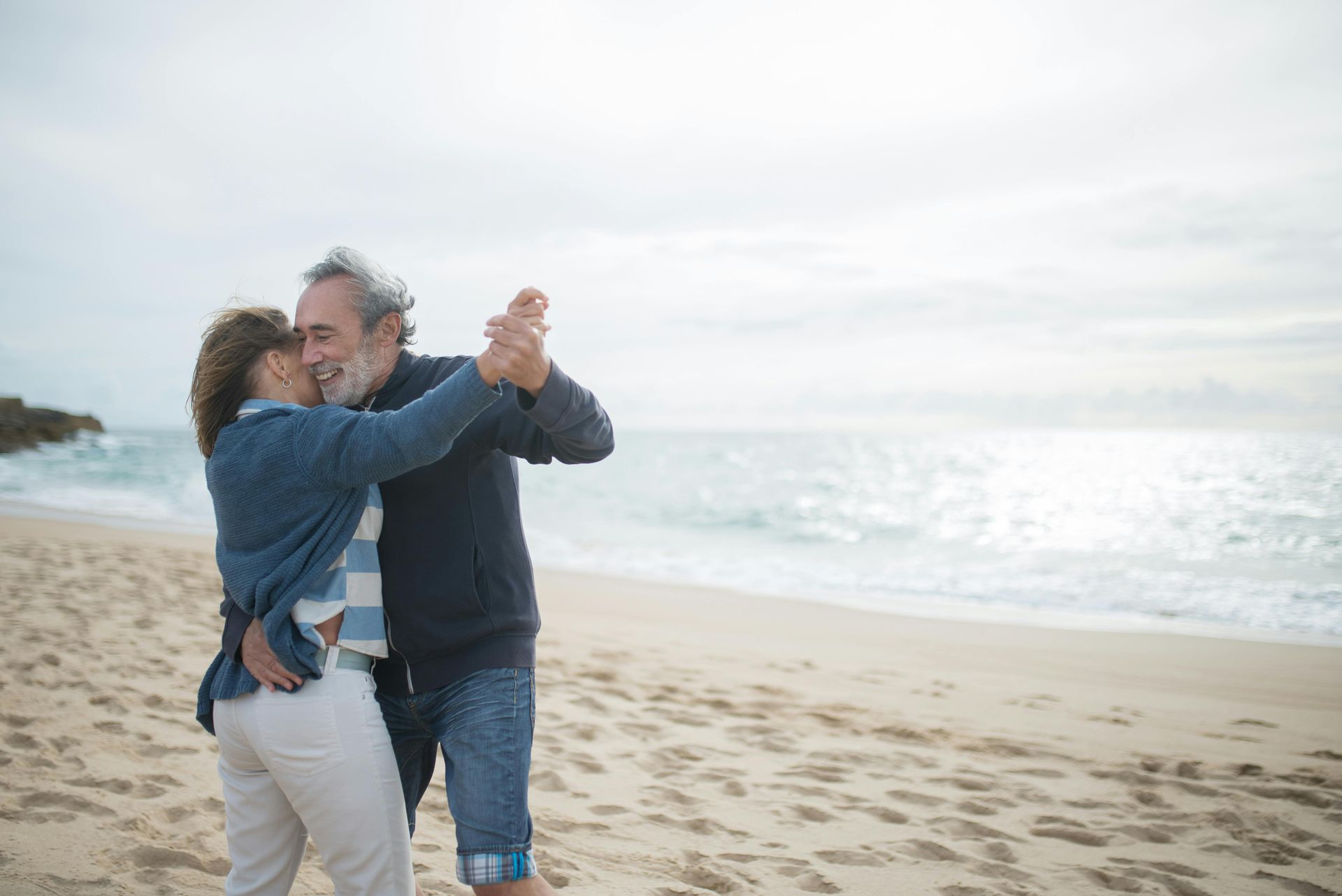 Elderly couple dancing joyfully on a sandy beach, ocean waves in the background.
