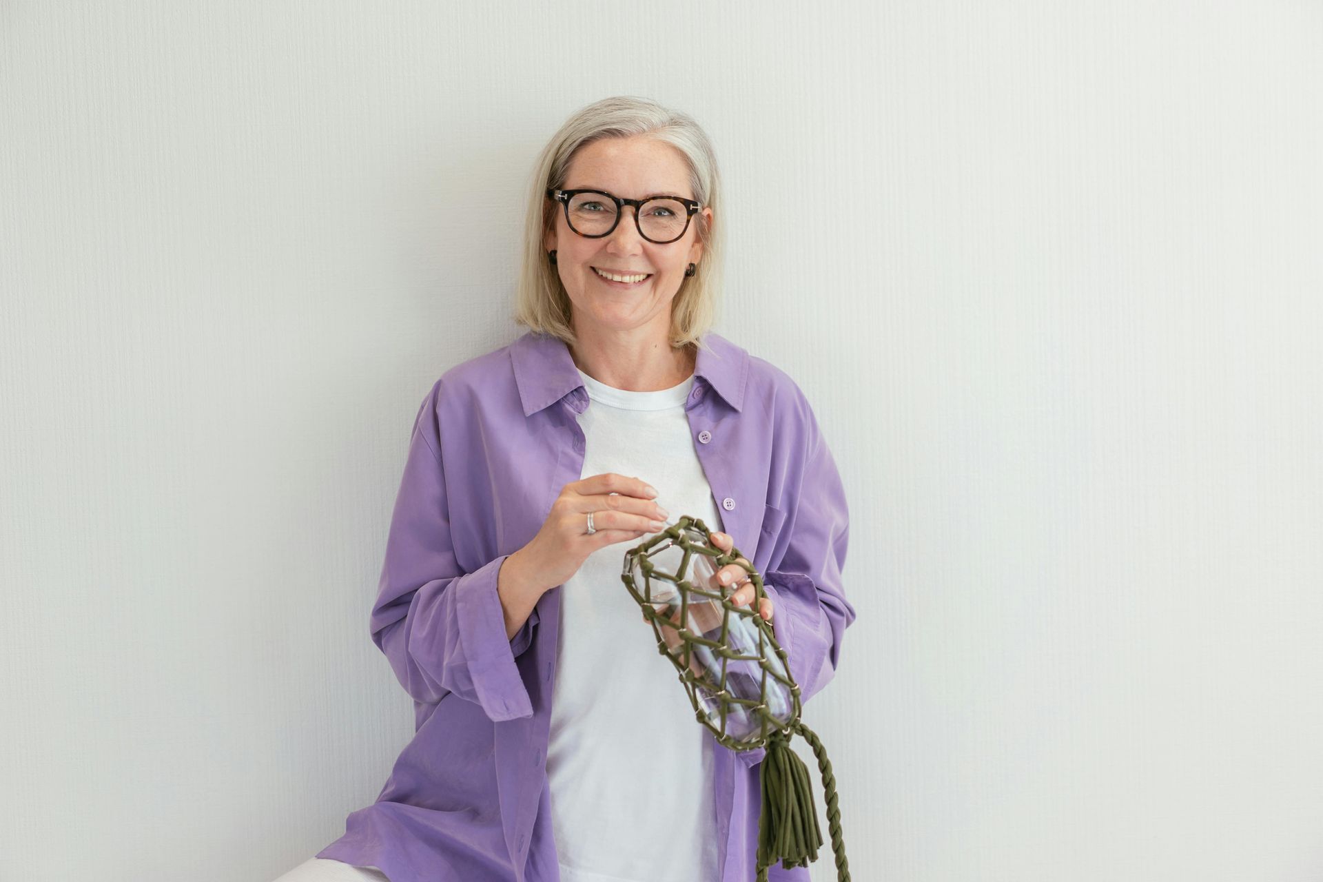 Woman with glasses smiles, holding a green macrame water bottle holder, wearing a purple shirt and white shirt.