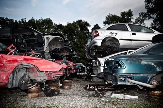 Stacked wrecked cars in a junkyard, including red, white, teal, and black vehicles on cinder blocks.