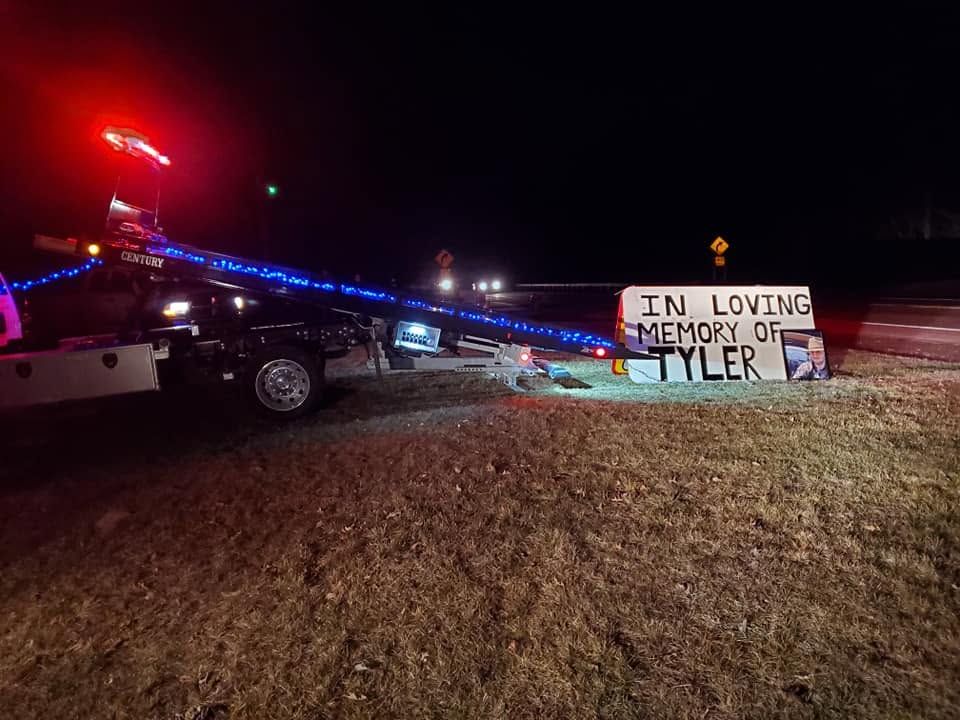 Tow truck at night near memorial sign reading “In loving memory of Tyler”