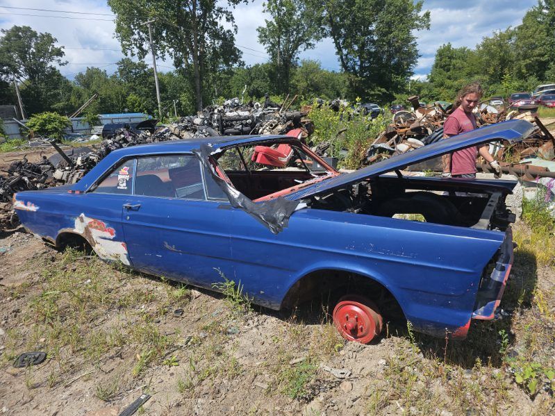Blue stripped car shell in a junkyard with missing windows and red wheels, surrounded by other wrecked cars.