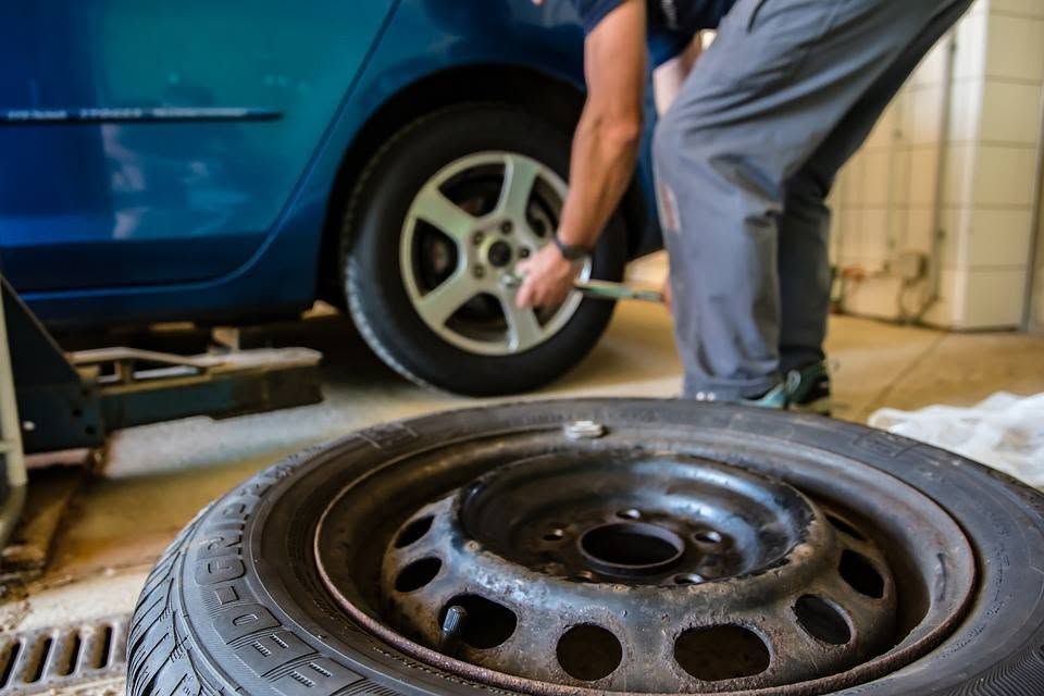 Person changing a car tire beside a blue vehicle in a garage, with a spare wheel in the foreground