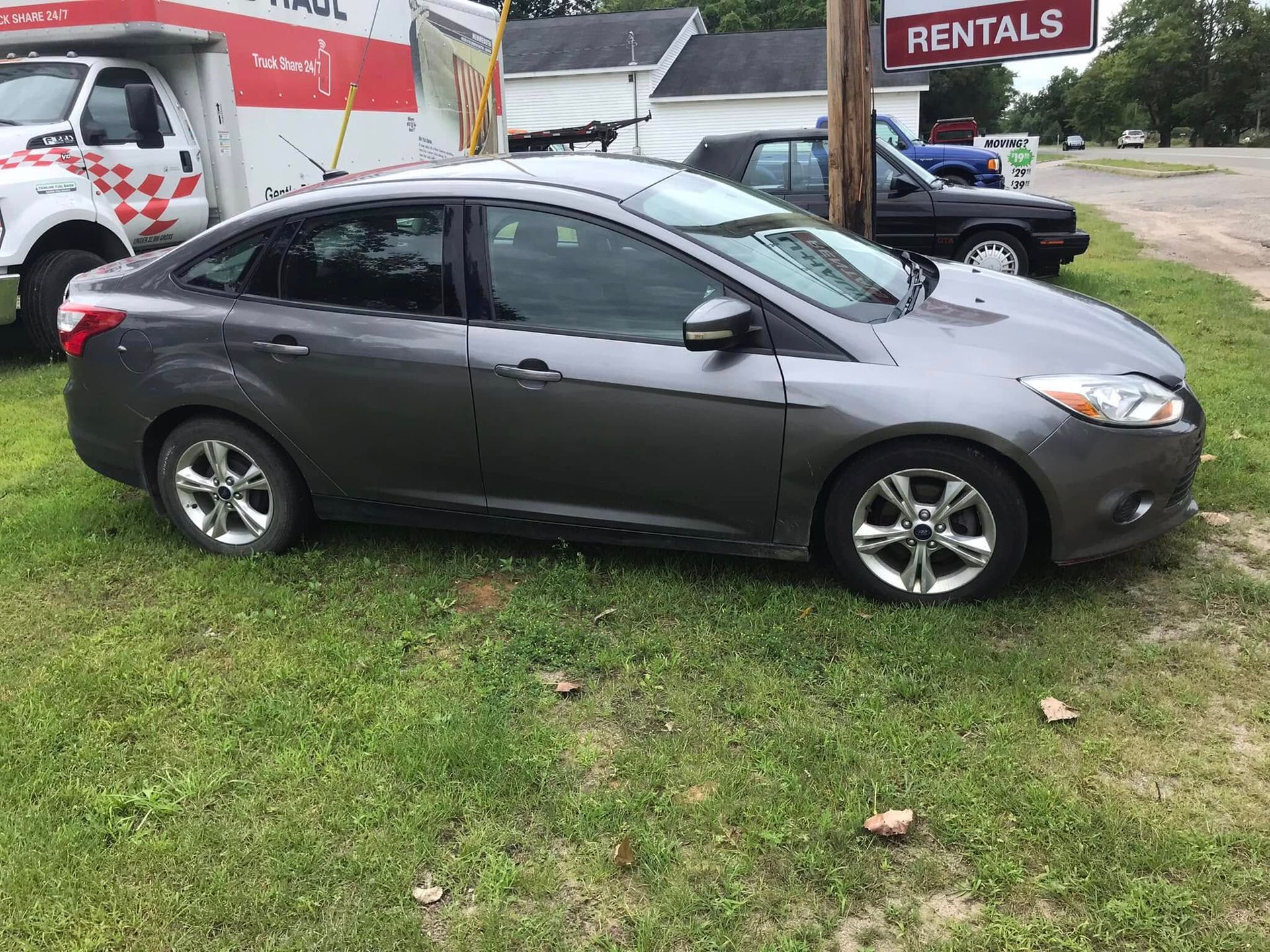 Gray sedan parked on grass beside a rental lot, with a red-and-white rental van in the background
