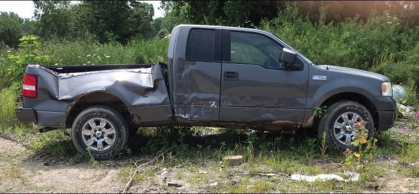 Damaged dark pickup truck with crushed rear side, parked in overgrown grass