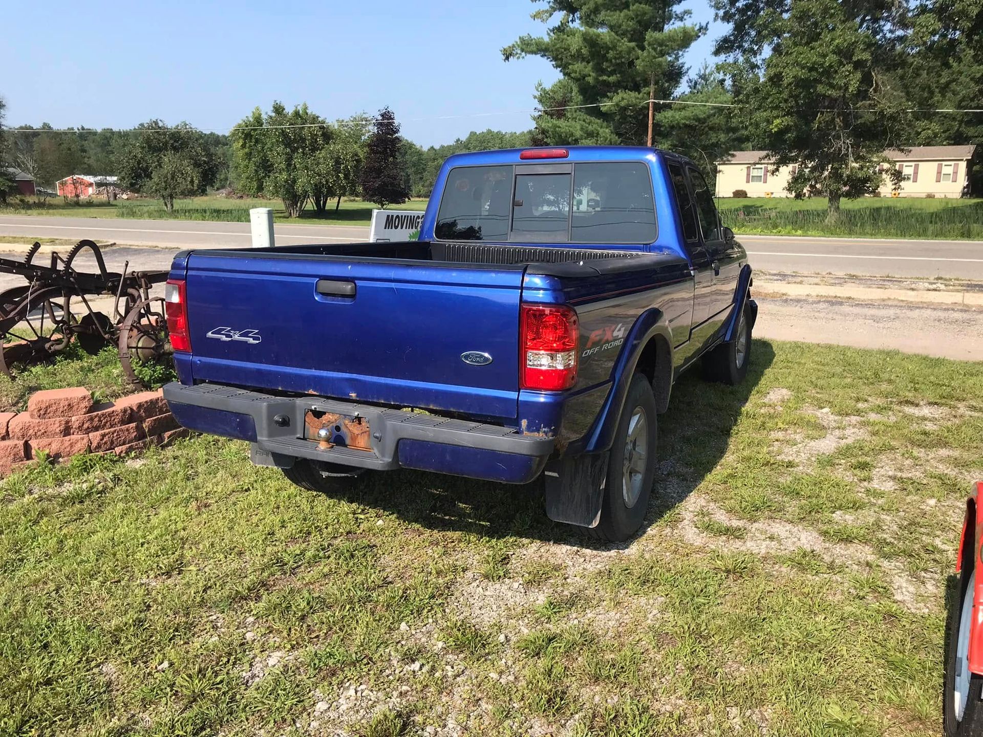 Blue pickup truck parked on grass beside a driveway, with trees and houses in the background