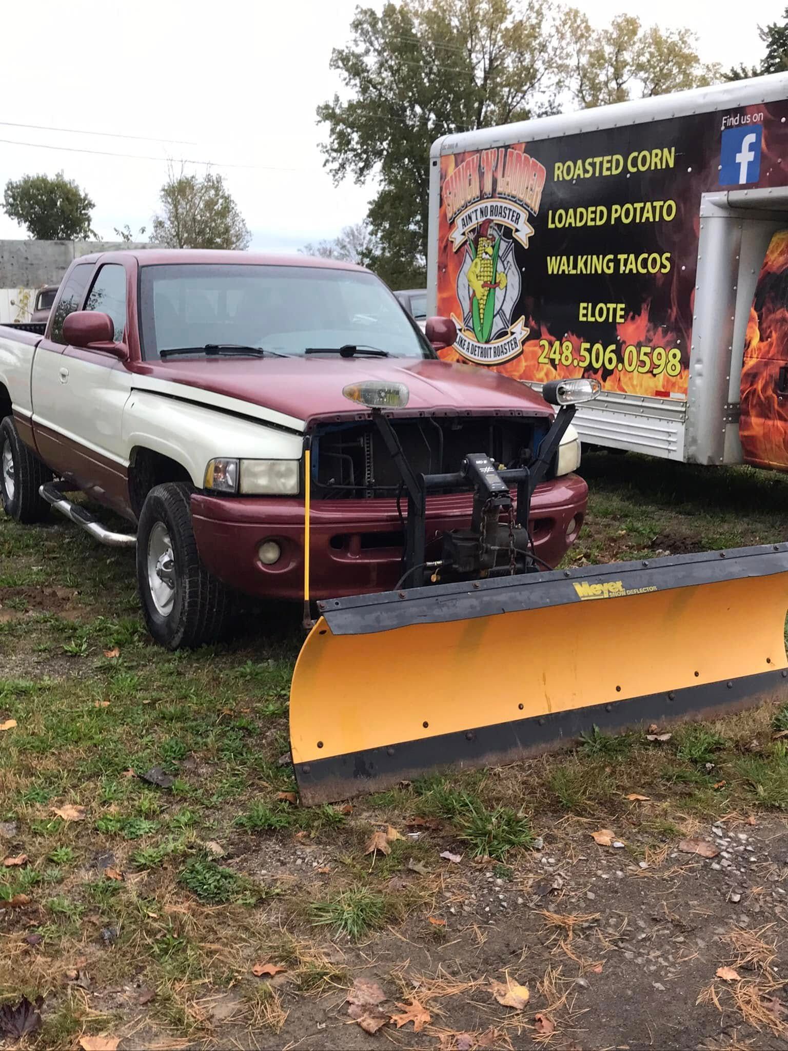 Maroon and cream vintage truck with plow in wet yard beside a colorful mural trailer.