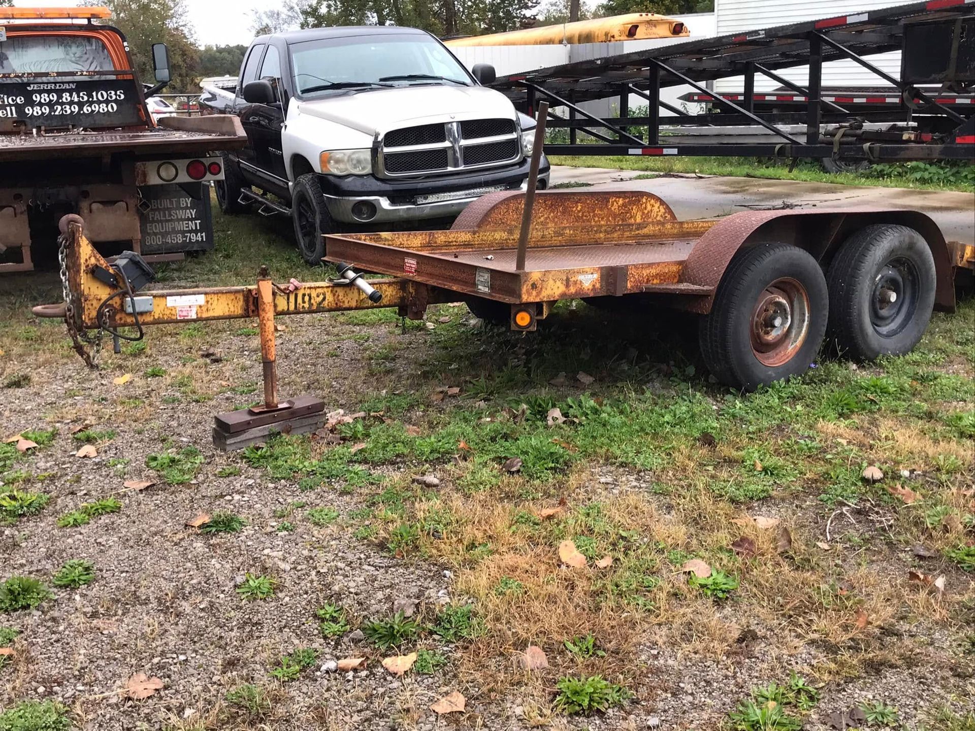 Rusty flatbed trailer with dual wheels parked on gravel beside a white pickup truck.