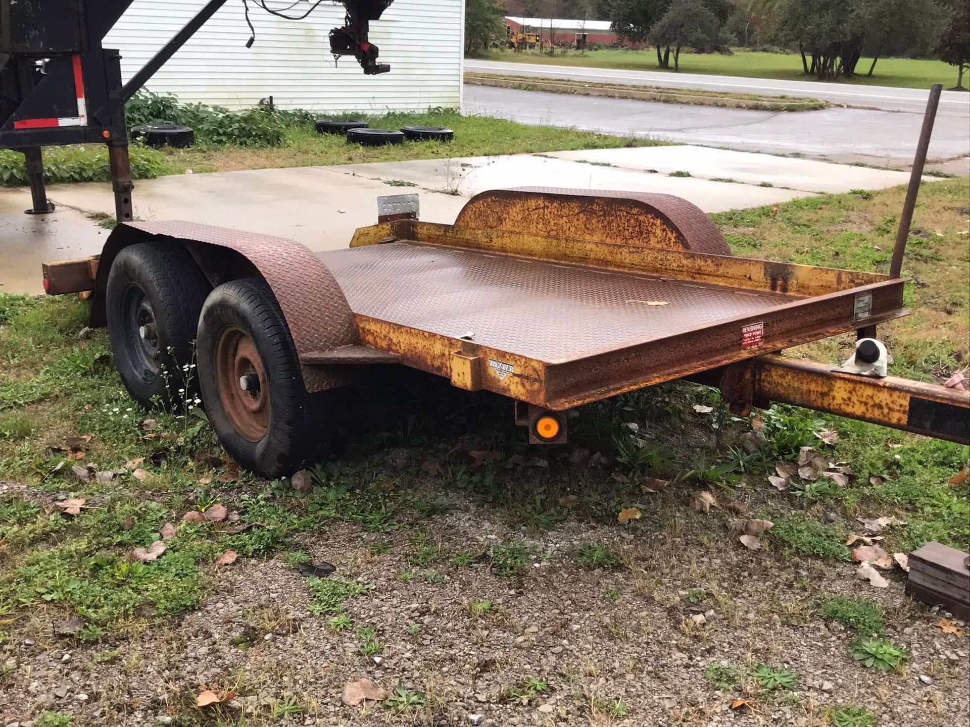 Rusty flatbed trailer with dual axles parked on grass beside a road