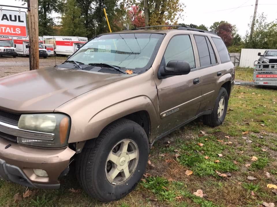 Brown SUV parked on grass by a roadside, with a dented front bumper and fall trees in the background