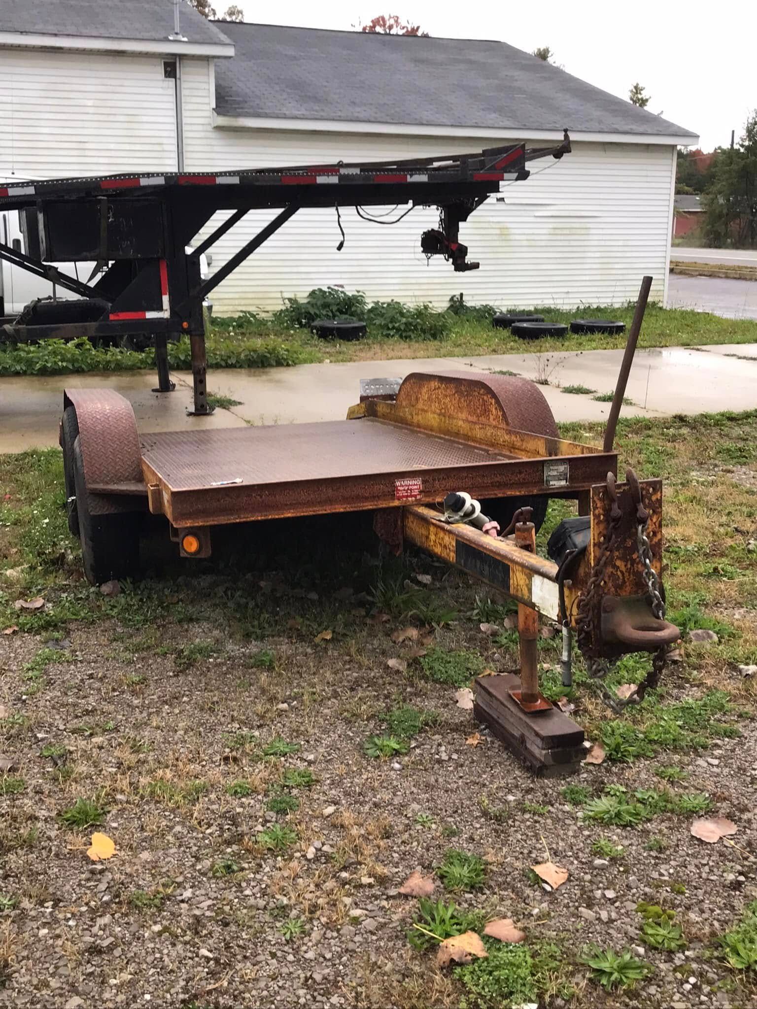 Rusty trailer frame and machinery parts on gravel beside a house and detached garage.