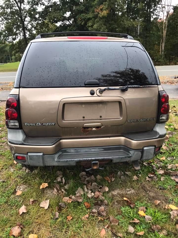 Rear view of a bronze SUV parked on grass, with rain on the rear window and a missing license plate.