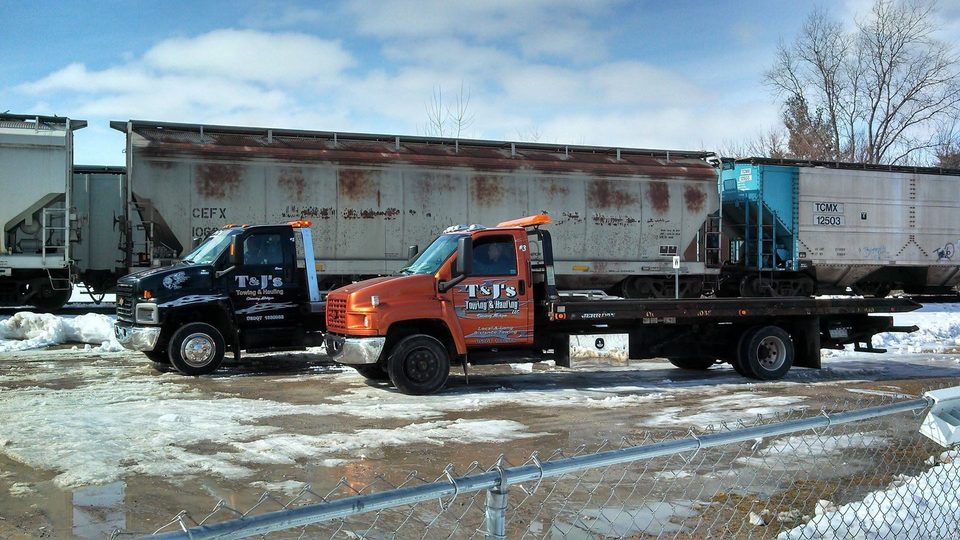 Two tow trucks parked in a snowy lot beside a weathered white trailer and other trucks.