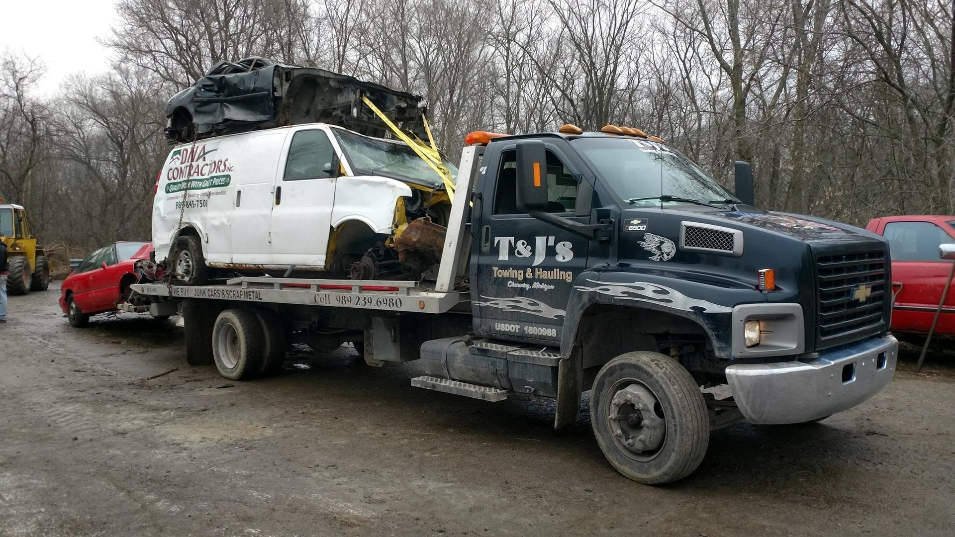 Tow truck carrying a damaged white van on a flatbed, parked on a dirt roadside with bare trees.