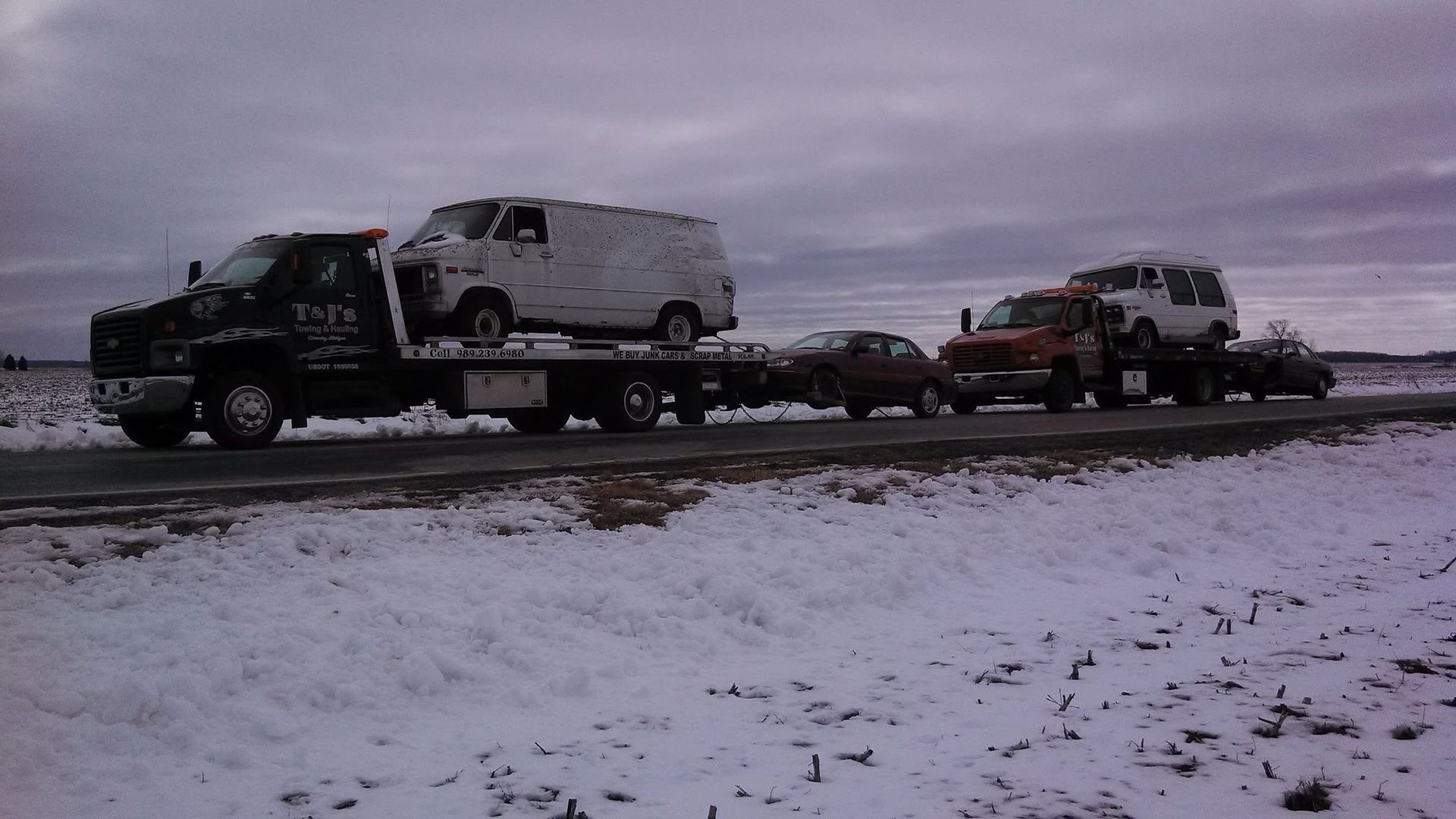 Tow trucks hauling damaged vehicles on a snowy roadside under a gray sky