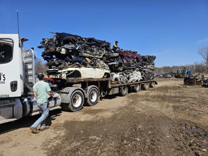 Loaded scrap metal truck at a muddy yard with a man standing beside it under a blue sky