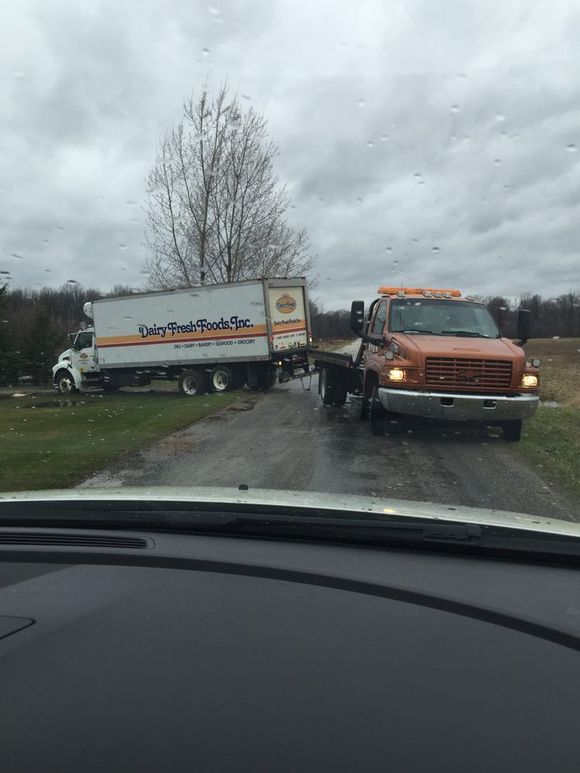 Orange tow truck and white box truck blocking a wet roadside under cloudy skies.