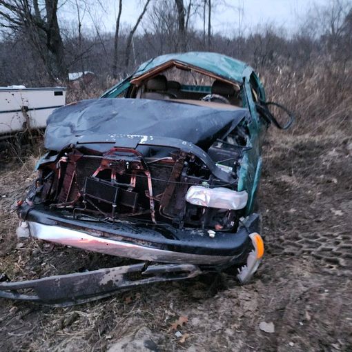 Wrecked black pickup truck off-road in brush, front end crushed and hood bent upward at dusk.