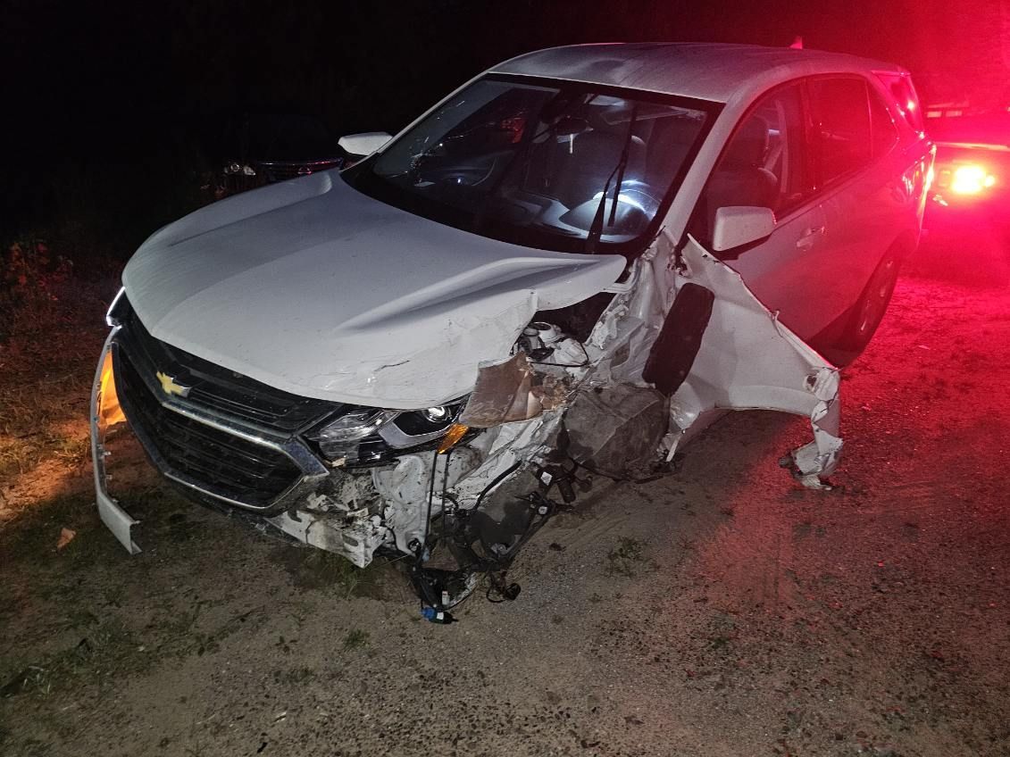 Damaged white car with front-end crash damage on a dark roadside at night, red emergency lights nearby